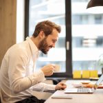 man-holding-teacup-infront-of-laptop-on-top-of-table-inside-925786