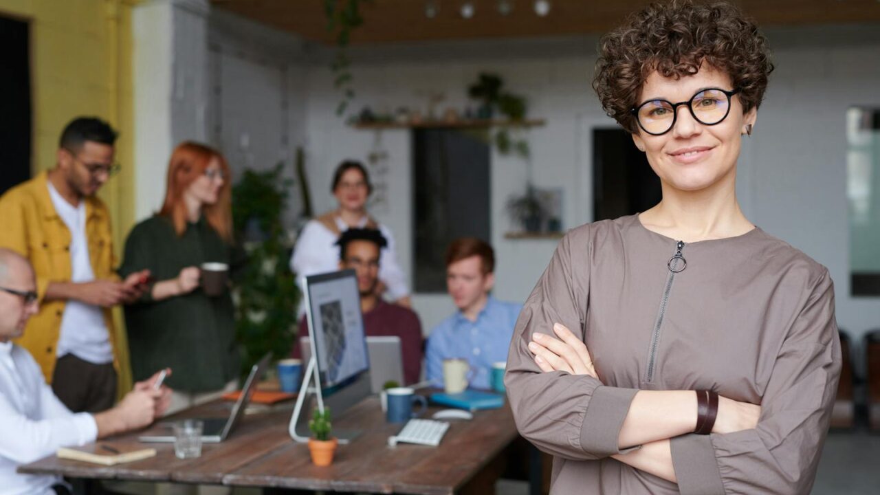 Une femme d'affaires souriante aux cheveux bouclés se tient avec confiance dans un espace de bureau moderne avec ses collègues.