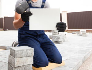 Artisan carreleur tenant un panneau blanc et un maillet lors d'un chantier extérieur à Belley
